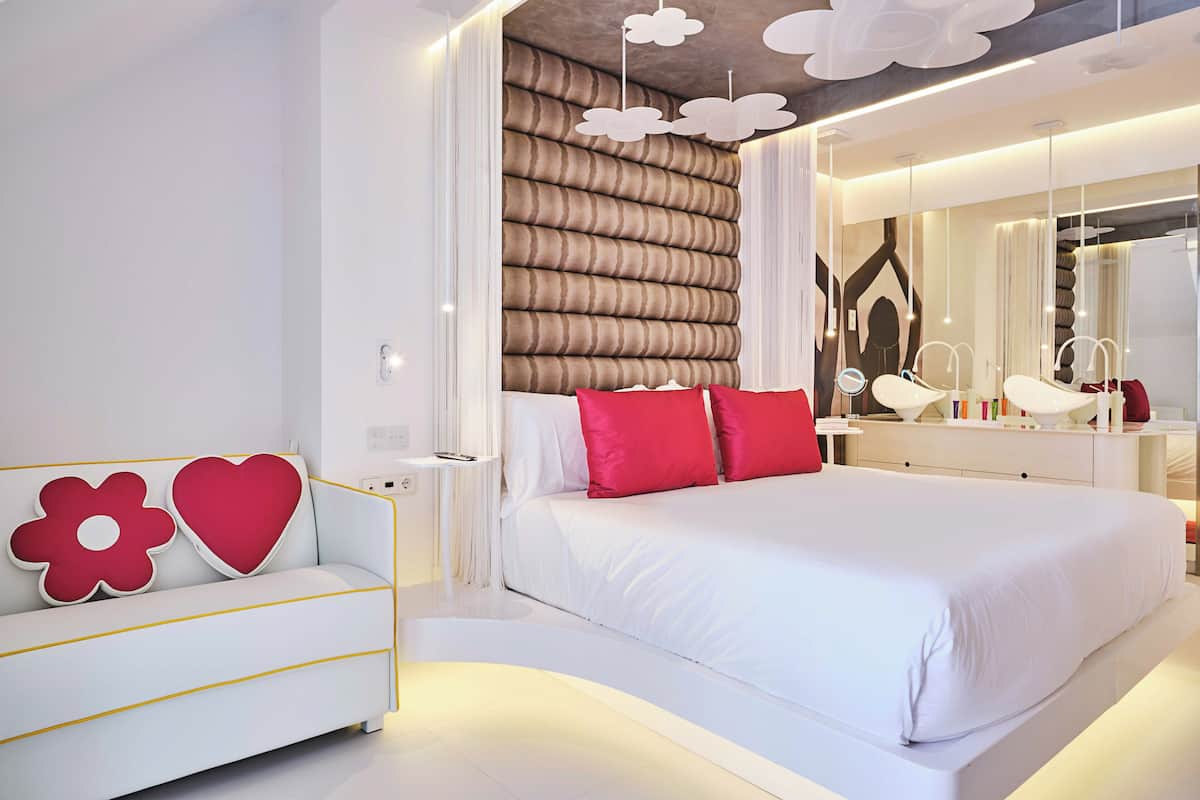 A modern bedroom with a large bed, a white sofa with red and white cushions, and a wall-mounted vanity with mirrors.
