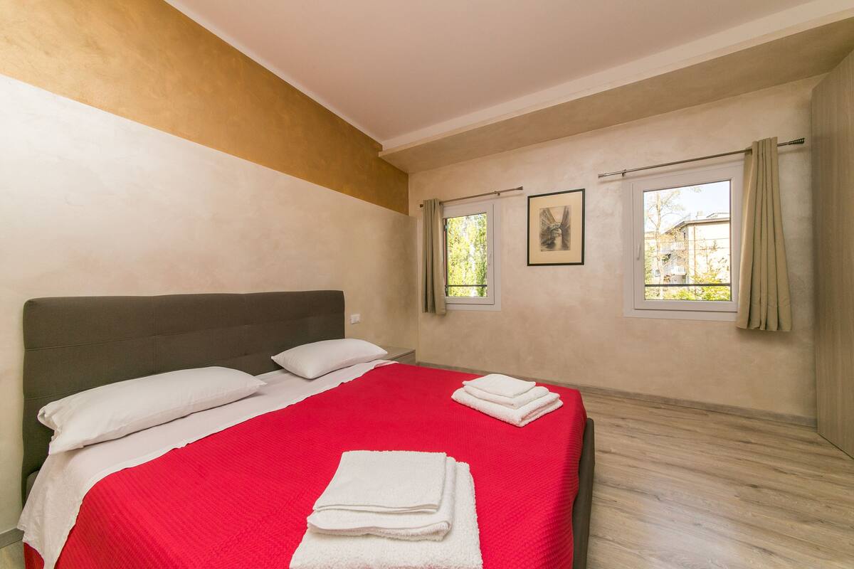 A bedroom with a large bed, two windows, and a view of the outside.