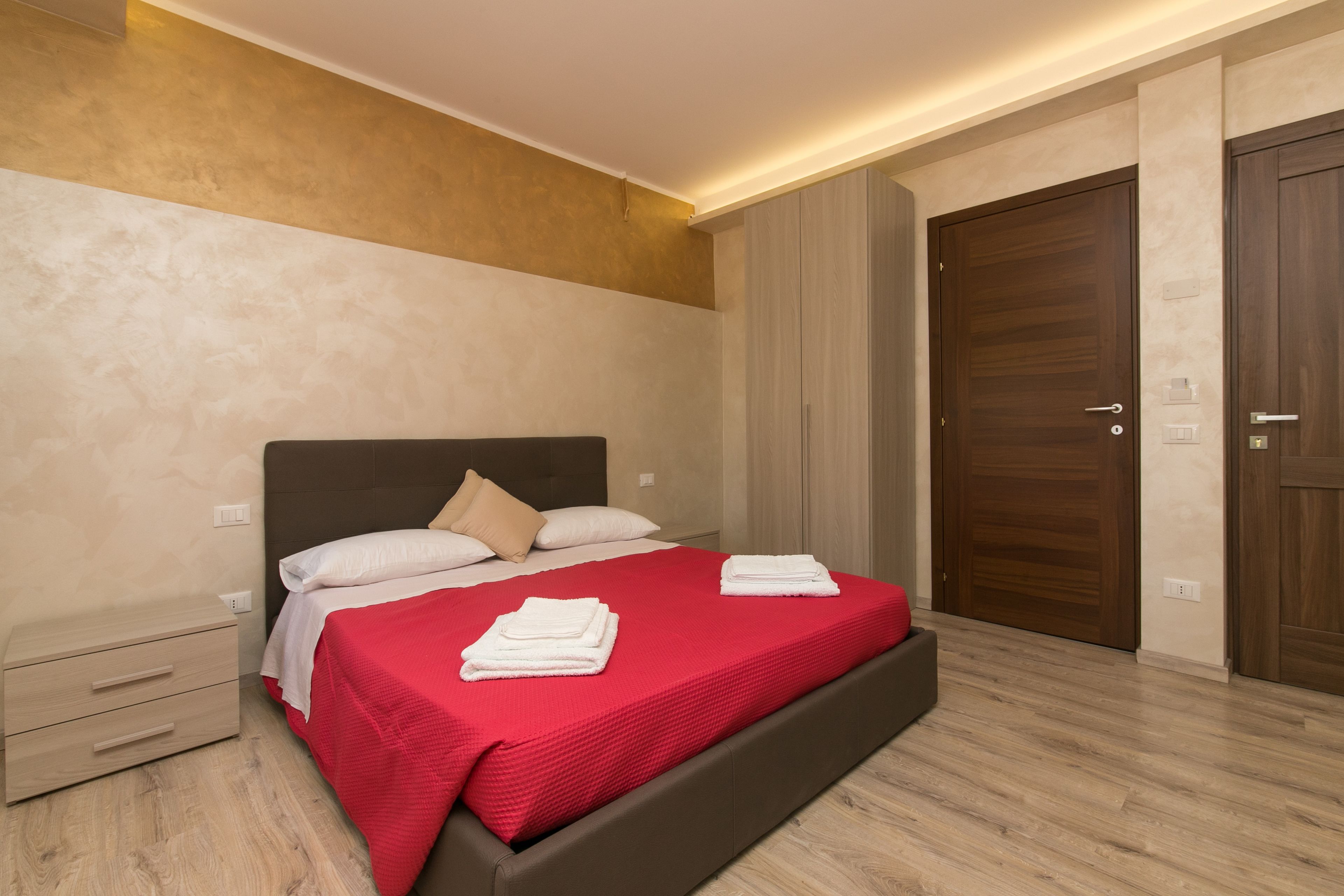 Una cama bien hecha con colcha roja y almohadas blancas, una mesita de noche de madera con cajones y una puerta de madera a la derecha.