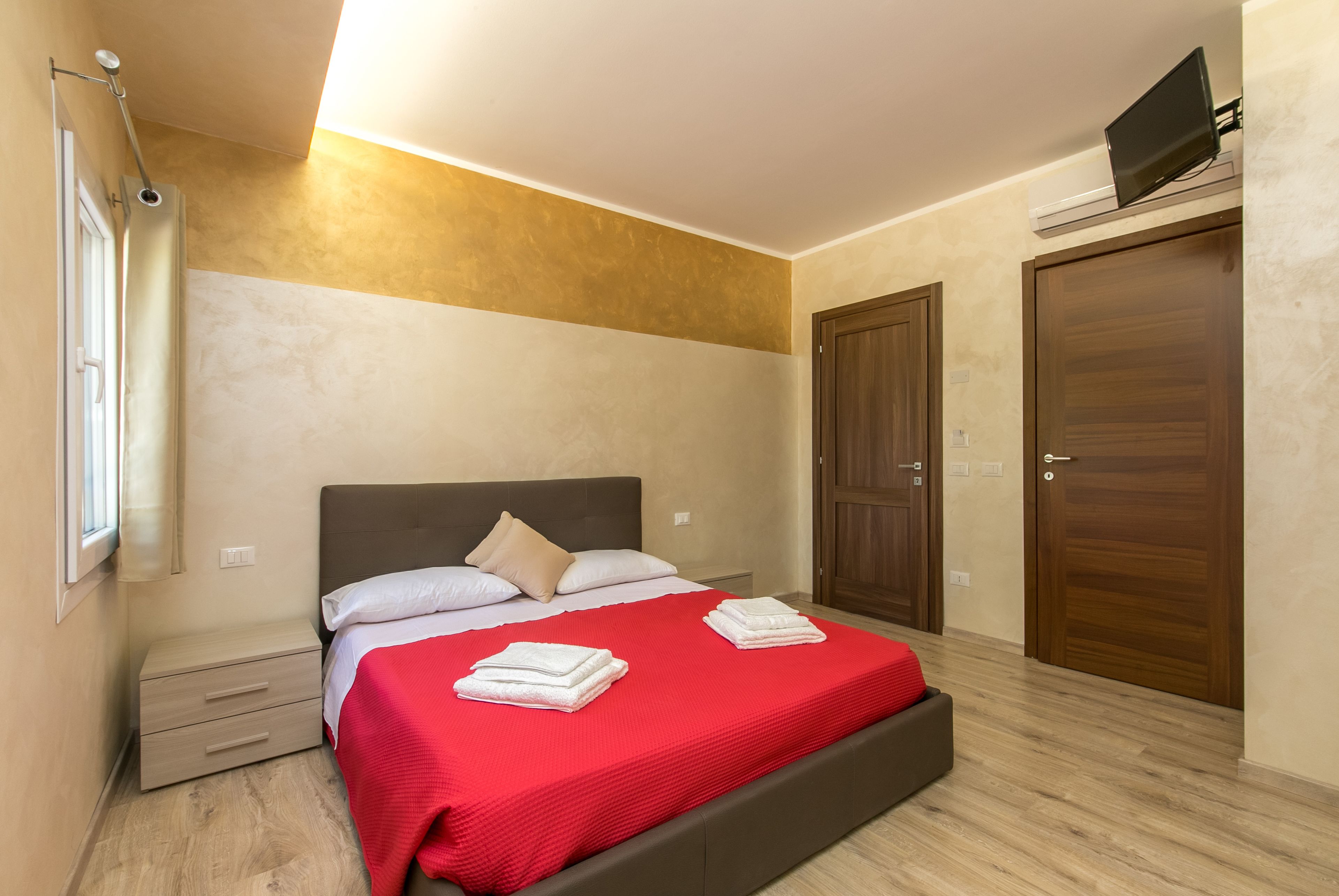 A bedroom with a bed, a nightstand, a red bedspread, a wooden wardrobe, and a TV on the wall.