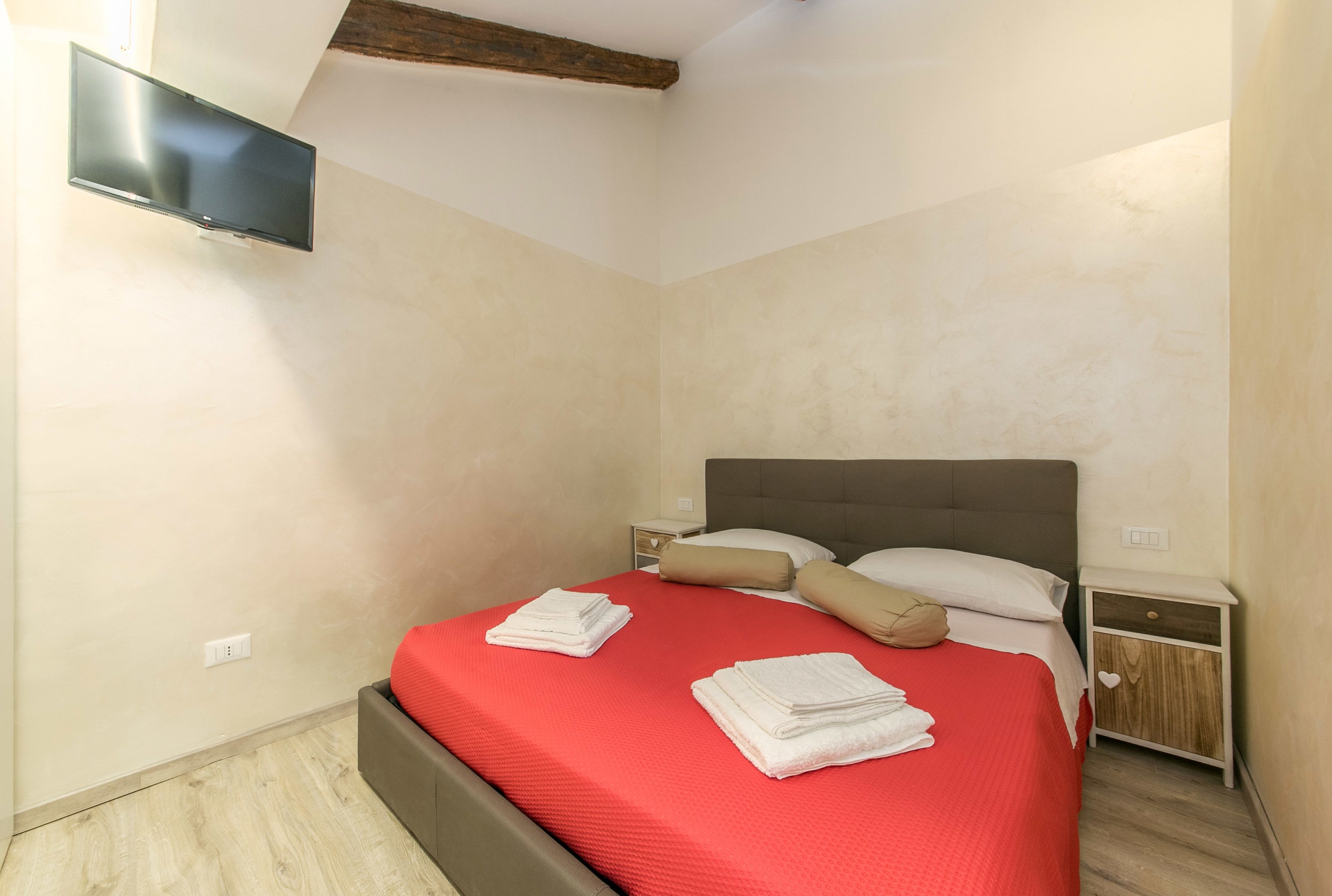 Un letto rifatto con copriletto rosso, due asciugamani bianchi piegati e un comodino in legno con un cassetto.