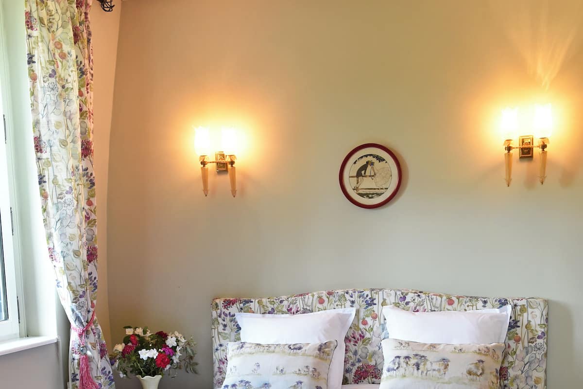 Una cama con cabecera floral, dos apliques, un reloj y un florero con flores.