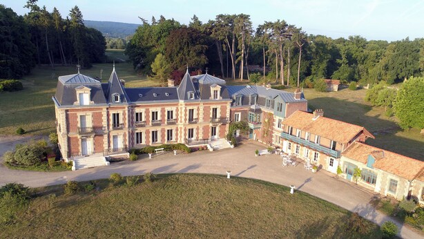 A large, multi-story building with a courtyard, surrounded by trees and a hilly landscape.