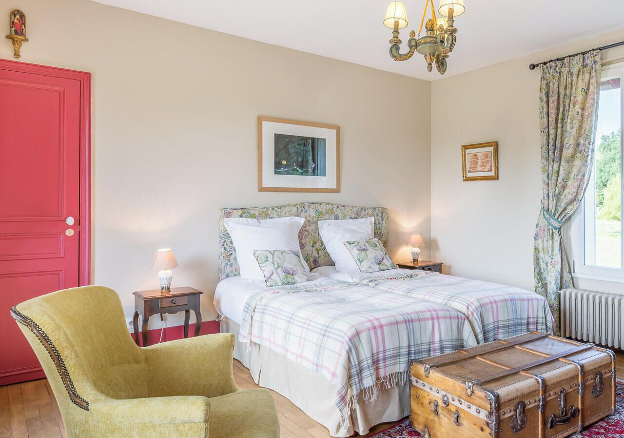 Een slaapkamer met een tweepersoonsbed, een stoel, een nachtkastje met een lamp, een koffer en een foto aan de muur.