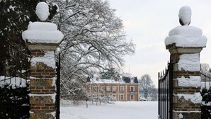 A snow-covered entrance with brick pillars and a gate, leading to a building with a snow-covered roof.
