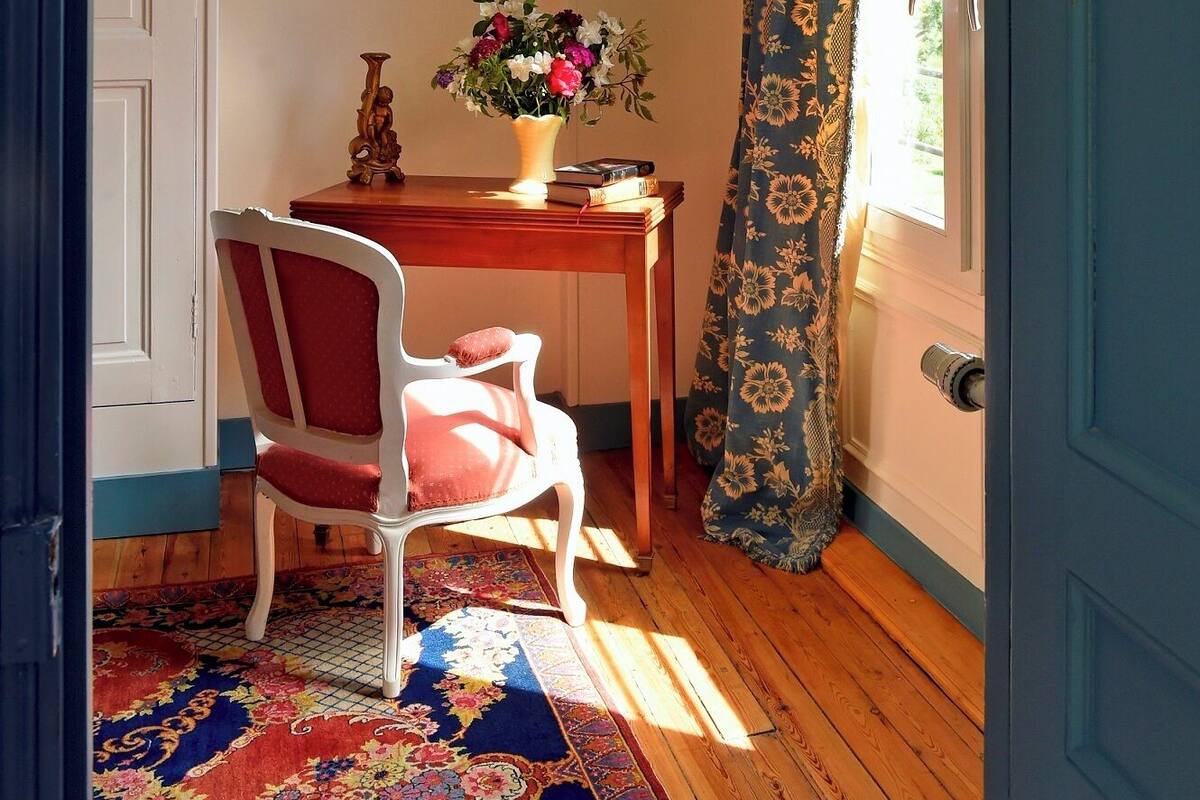Una habitación con piso de madera, una silla blanca, una mesa con un jarrón de flores, un cuadro en la pared y una puerta azul.