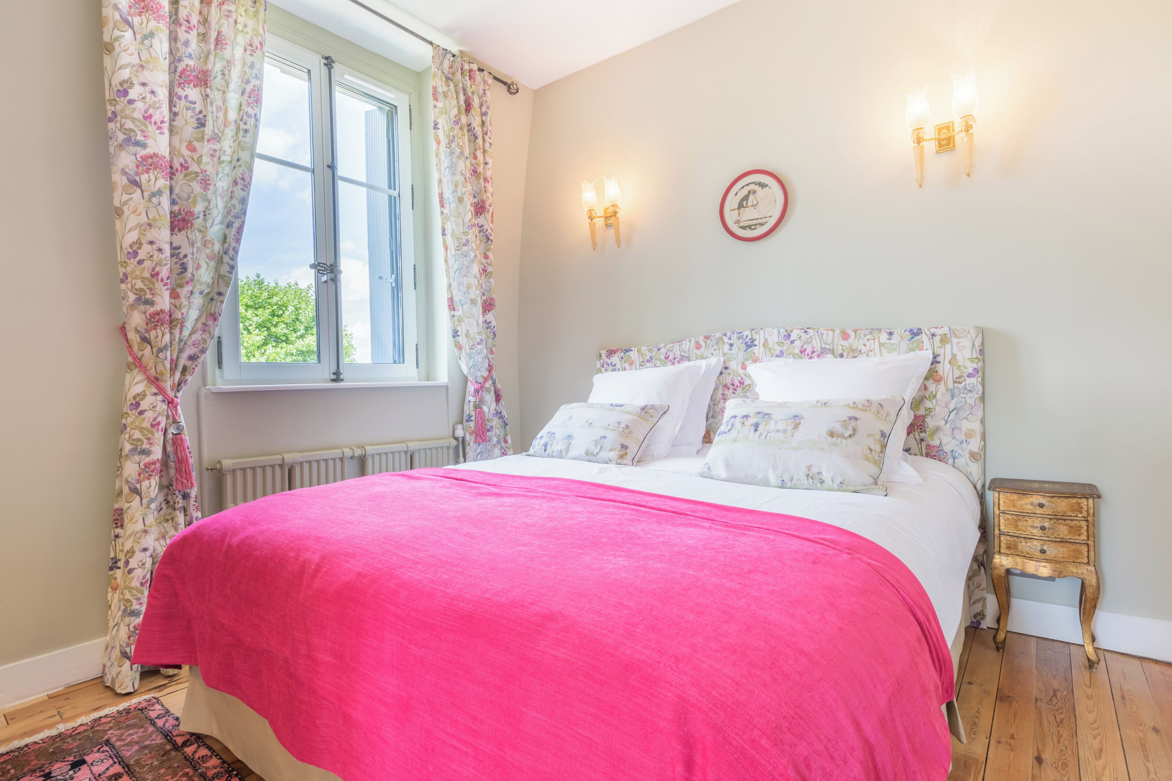 Een slaapkamer met een bed, bloemen gordijnen, een nachtkastje en een wandklok.
