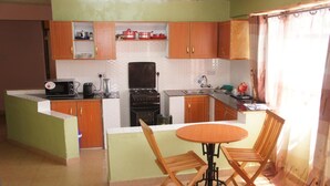 Een kleine keuken met een gasfornuis, magnetron en houten kastjes. Er is een eethoekje met een tafel en twee stoelen.