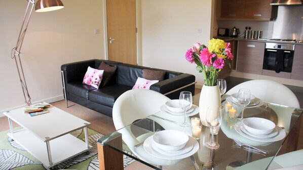 A dining area with a glass table set for four, surrounded by white chairs. A vase with flowers is on the table, and there is a black leather sofa in the background.