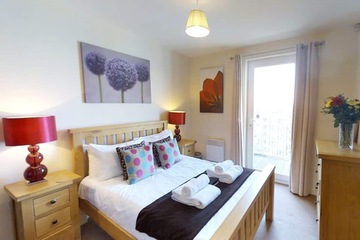 A bedroom with a wooden bed, two bedside tables, a nightstand, a window with curtains, and a view of buildings outside.