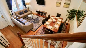 A spacious living area with a sofa, coffee table, and dining table set for six. A TV is mounted on the wall, and there are two framed artworks above the table.