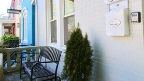 A stone-floored porch with a bench and a potted plant, a white wall with a mailbox and a number sign, and a black lantern.