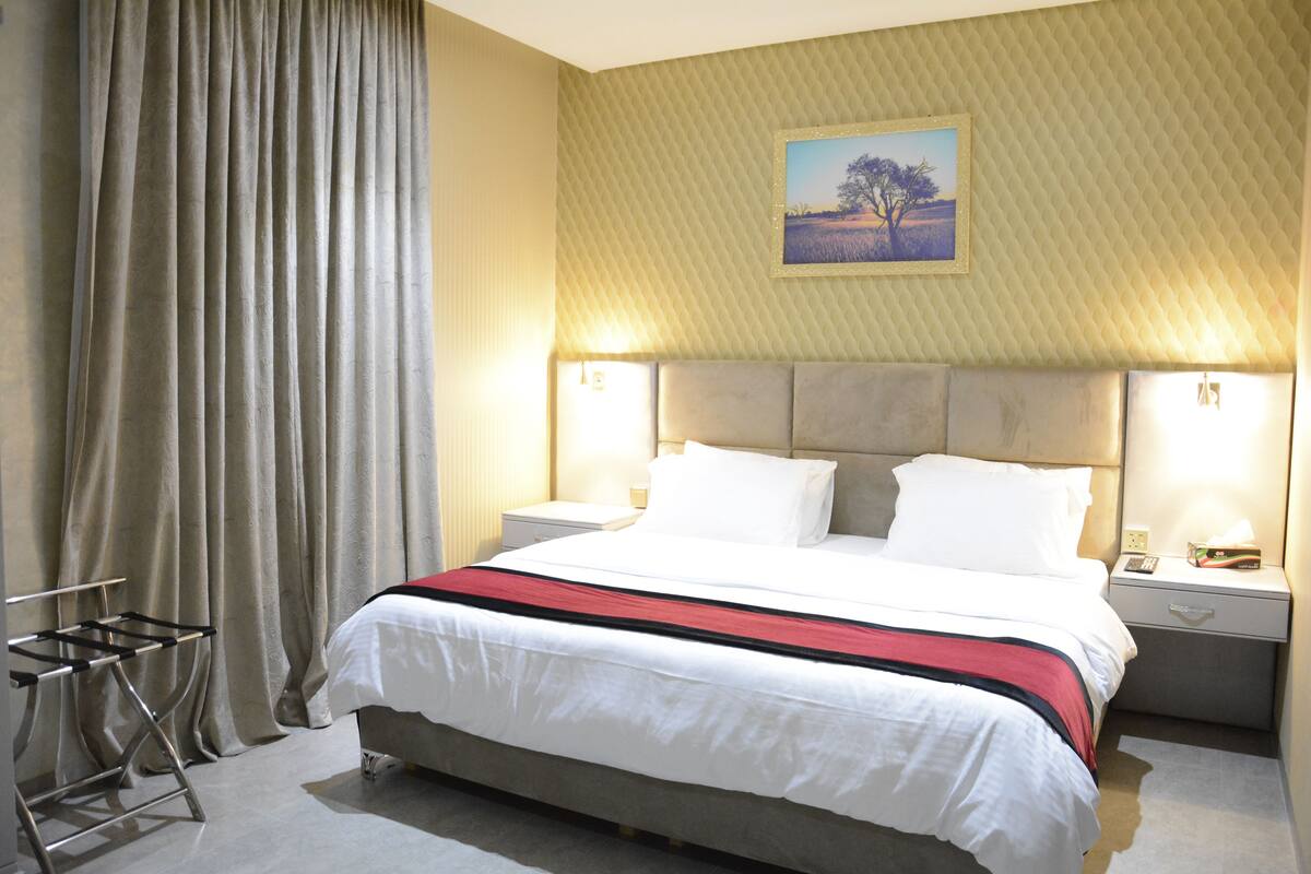 Ein Hotelzimmer mit einem großen Bett, zwei Nachttischen mit Lampen, einem Fenster mit Vorhängen und einem Gemälde an der Wand.