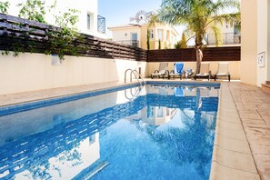 A swimming pool with a wooden deck and lounge chairs, surrounded by a low wall and a palm tree. - Natalie Villa CL 41 (Paralimni)