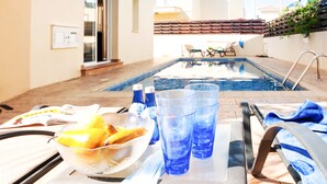Escena junto a la piscina con una mesa, sillas, un cuenco de fruta y vasos azules.