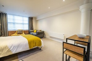 A modern hotel room with a bed, a small sofa, a table with a bowl, and a window with curtains.