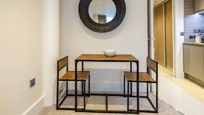 Uma área de refeições com uma mesa de madeira e duas cadeiras, um espelho redondo na parede e uma cozinha visível através de uma porta aberta.