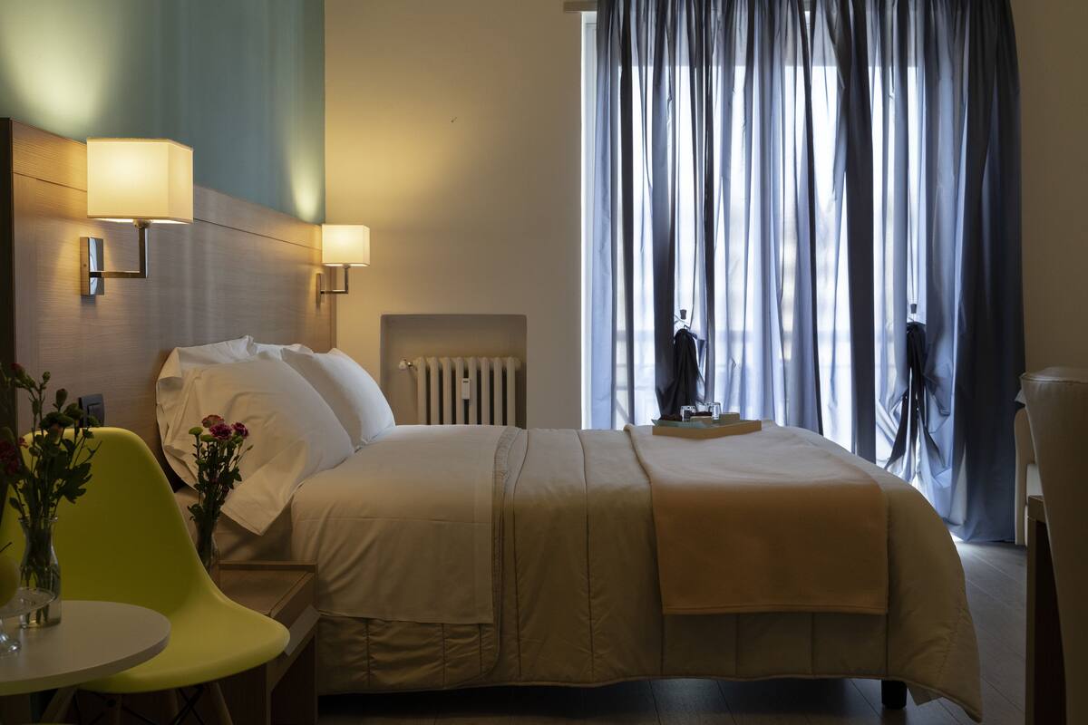 A hotel room with a bed, bedside lamps, a radiator, a chair, and a window with curtains.