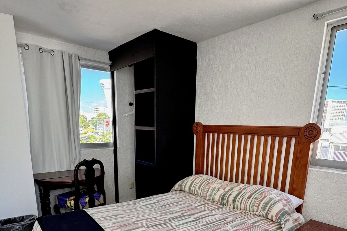 Une chambre à coucher avec un lit, une tête de lit en bois, une fenêtre donnant sur un beau paysage, une petite table et une poubelle.
