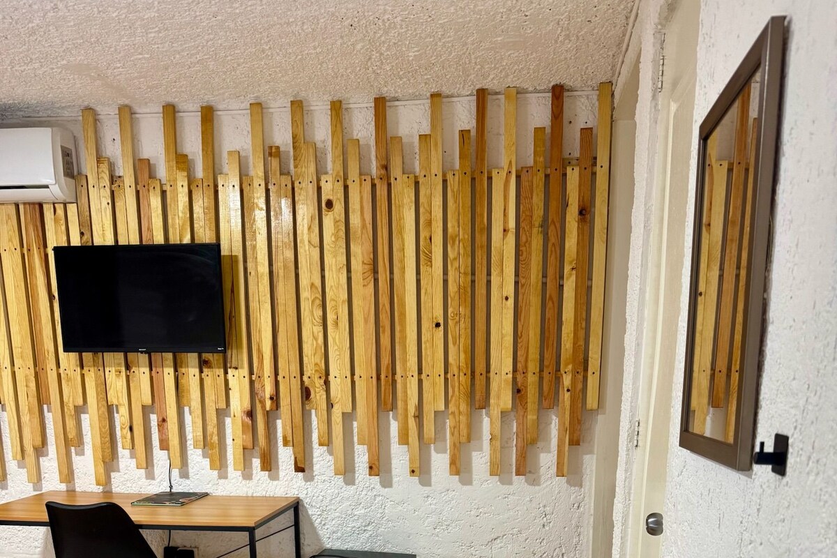 Een kamer met een wand van houten latten, een flatscreen-tv, een bureau met stoel, een kleine koelkast, een wastafel en een bed met een decoratieve spreidekking.