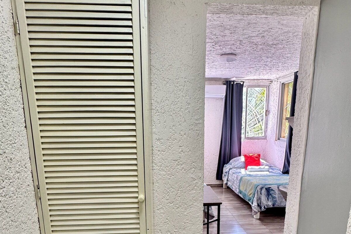 Un corridoio stretto che conduce a una camera da letto con un letto, un tavolino e una porta con persiane orizzontali.