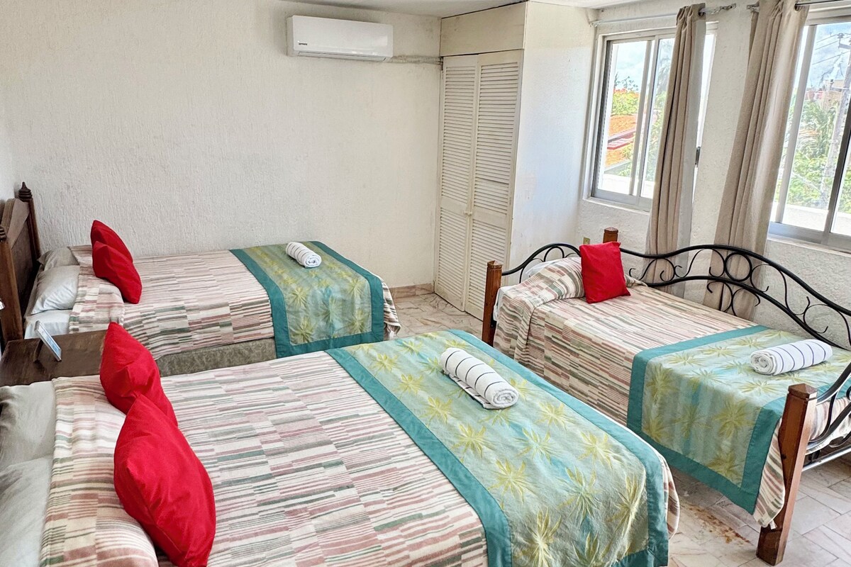 Camera d'albergo con tre letti, ognuno con copripiumino a righe e cuscini rossi, una finestra con tende e un condizionatore d'aria a parete.