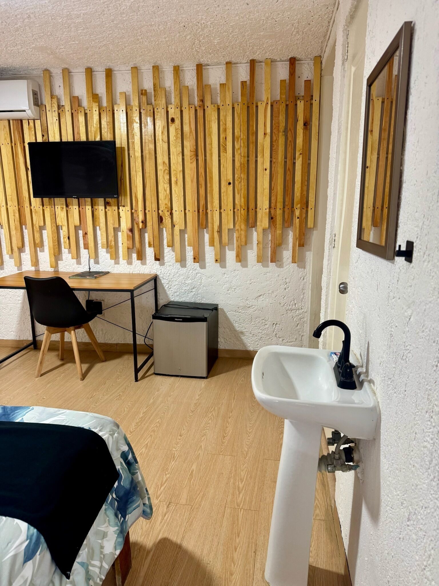 Une chambre avec un mur recouvert de lambris en bois, un téléviseur à écran plat, un bureau avec une chaise, un petit réfrigérateur, un évier et un lit recouvert d’une couverture à motifs.