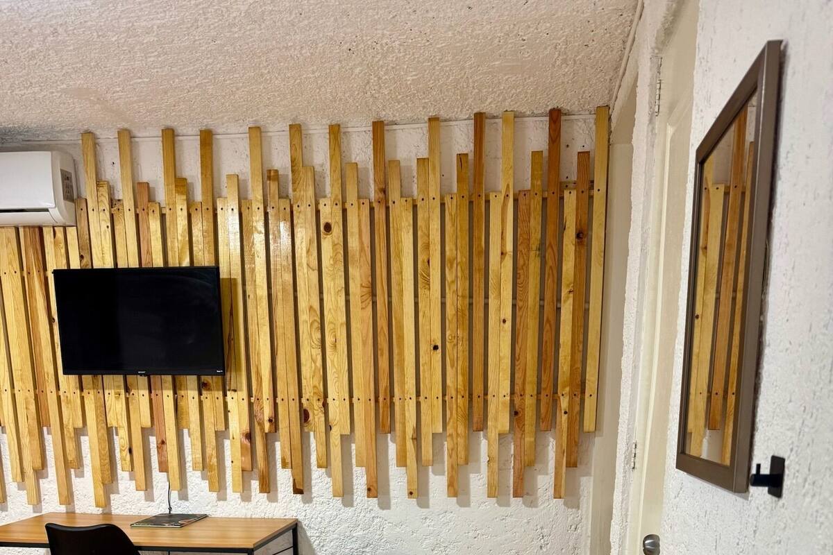 A room with a wooden slat wall, a flat-screen TV, a desk with a chair, a small refrigerator, a sink, and a bed with a patterned cover.