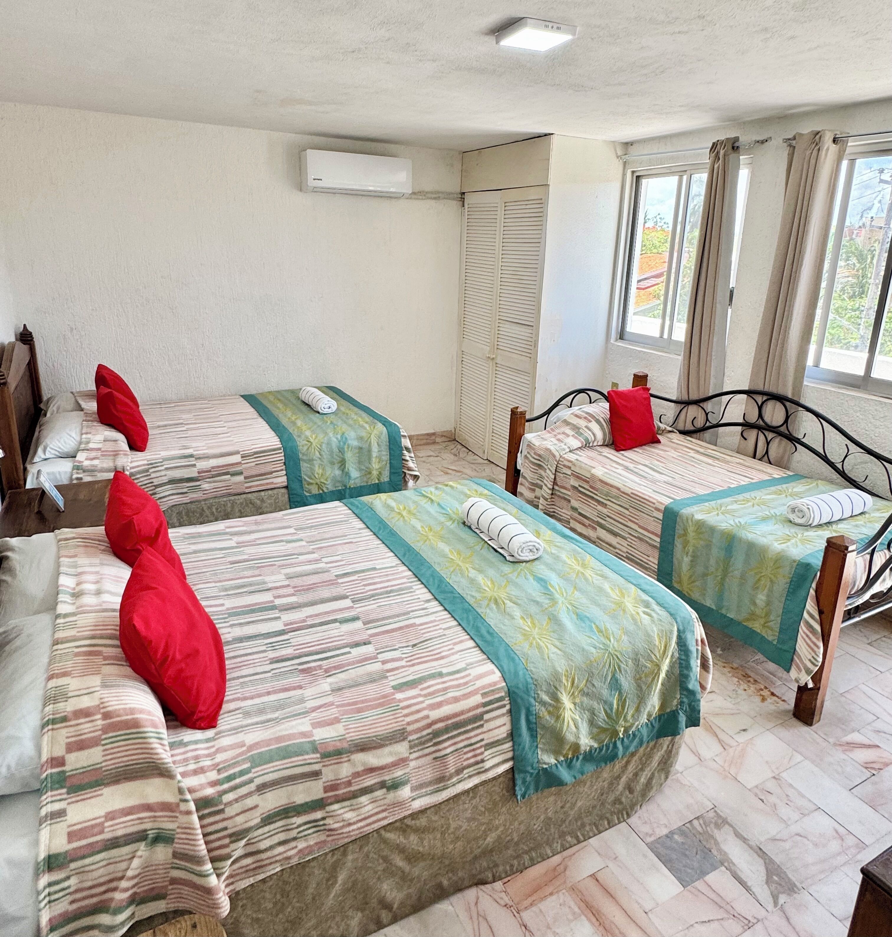 Une chambre d’hôtel avec trois lits, chacun recouvert d’une couvre-lit à rayures et agrémenté d’oreillers rouges, une fenêtre avec des rideaux et un climatiseur fixé au mur.