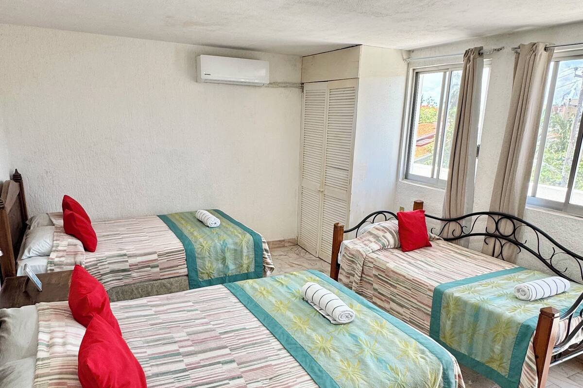 A hotel room with three beds, each with a striped bedspread and red pillows, a window with curtains, and an air conditioning unit on the wall.