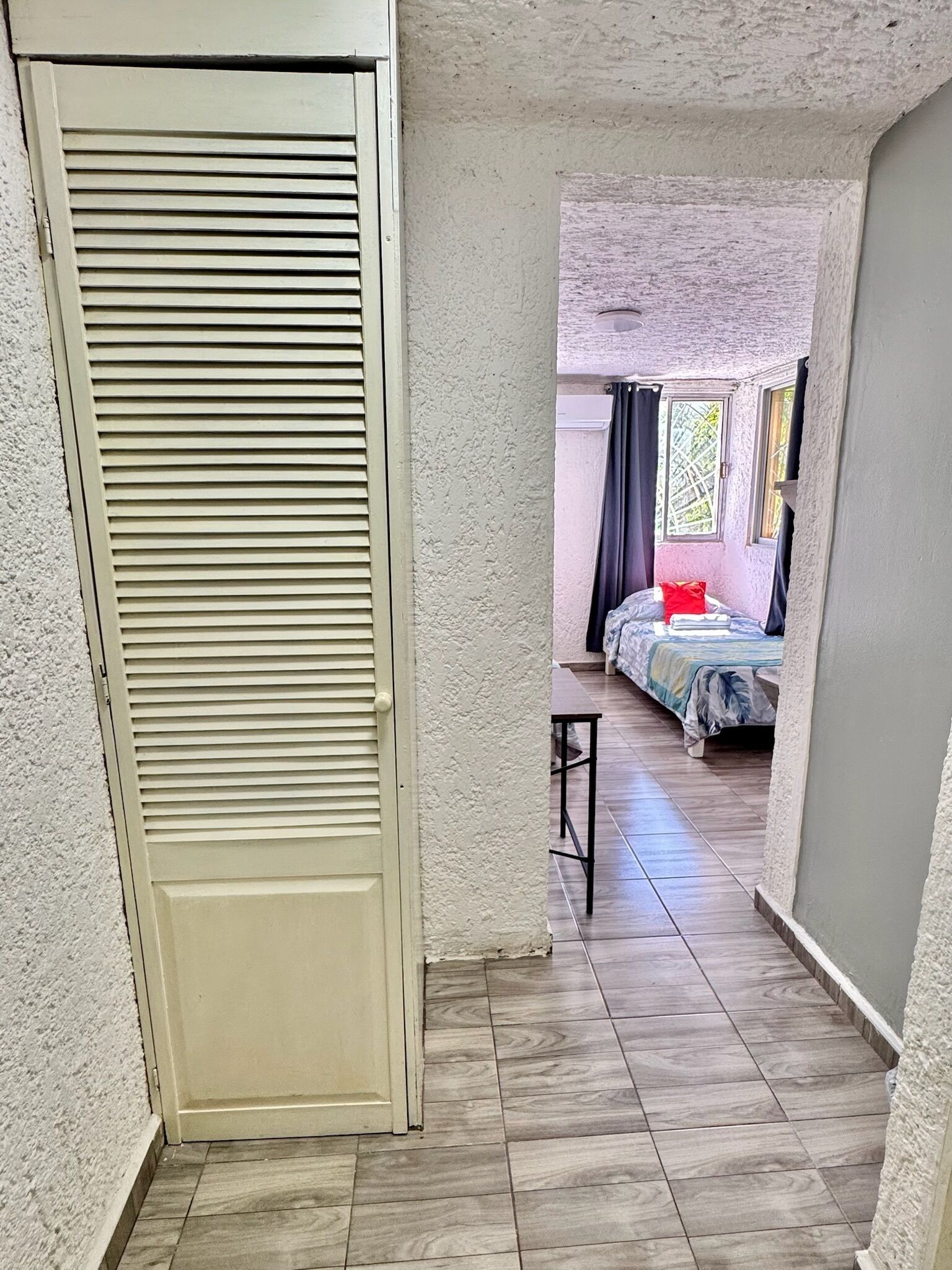 Un couloir étroit mène à une chambre qui contient un lit, une petite table et une porte à lattes horizontales.