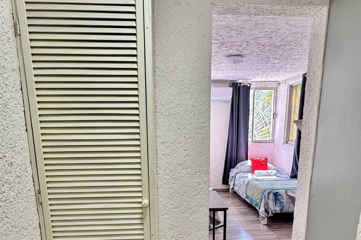 A narrow hallway leading to a bedroom with a bed, a small table, and a door with horizontal slats.