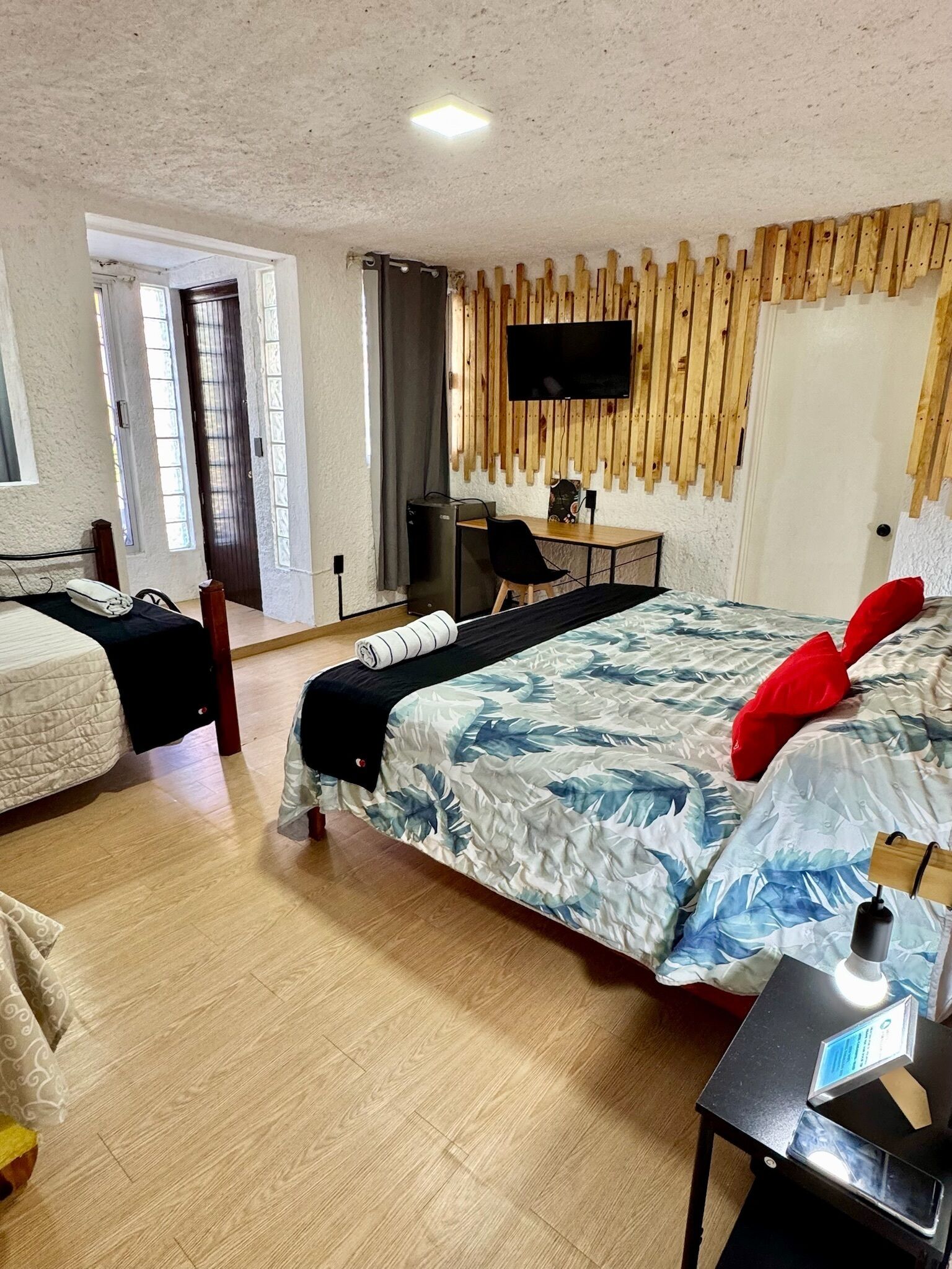 Une chambre avec un lit, un bureau, une chaise, un téléviseur fixé au mur (qui est en bois) et une porte donnant sur un balcon.