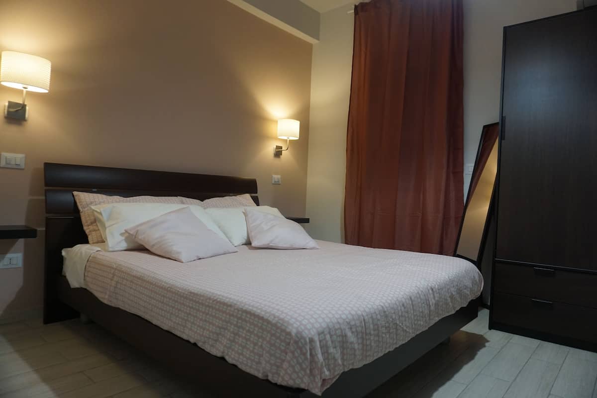 A neatly made bed with a headboard, two wall-mounted lamps, and a dark wardrobe.