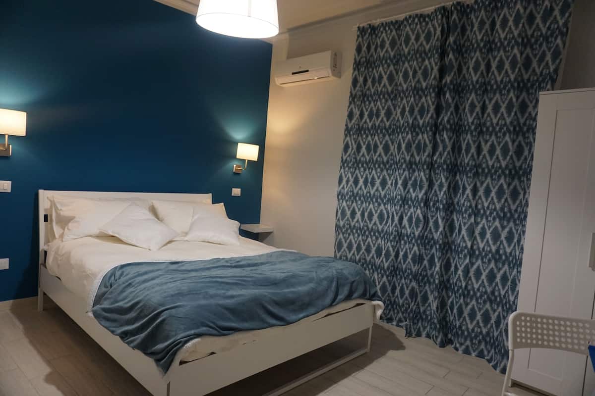A bedroom with a white bed, blue walls, patterned curtains, and a white chair.