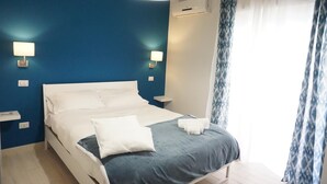 Una cama bien tendida con ropa de cama blanca y azul, dos lámparas de noche y una ventana con cortinas azules.