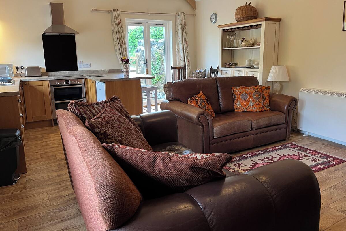 A cozy living room with a brown leather sofa, a kitchen area with a stove and oven, and a dining table.