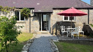 En stenbelagt hytte med en grussti, der fører til en terrasse med hvide metalstole og en rød parasol.
