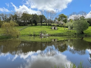 A serene pond with a wooden dock, surrounded by lush greenery and a small stone structure on a grassy hill. - Coombeshead Farm (Newton Abbot)