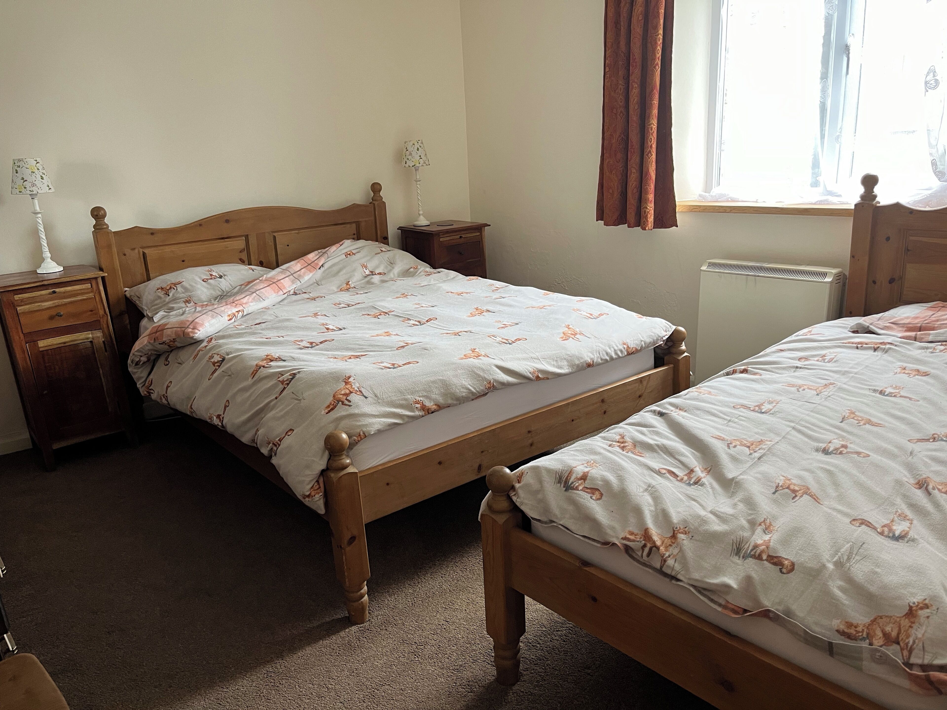 Quarto com duas camas de madeira, uma mesa de cabeceira com abajur, cômoda e janela com cortinas.