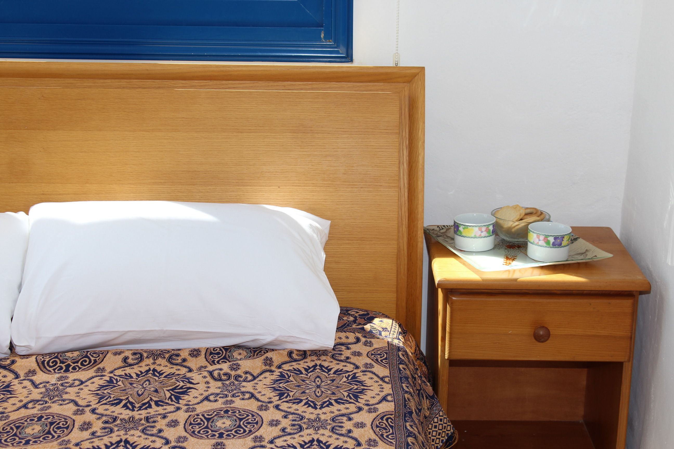 Een bed met een beddegoed met patroon, een houten hoofdbord en een nachtkastje met een lade en twee bekers.