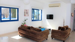 A living room with two brown sofas, a small table, a TV, and a window with blue frames. - Sirina Villa STN 29 (Paralimni)