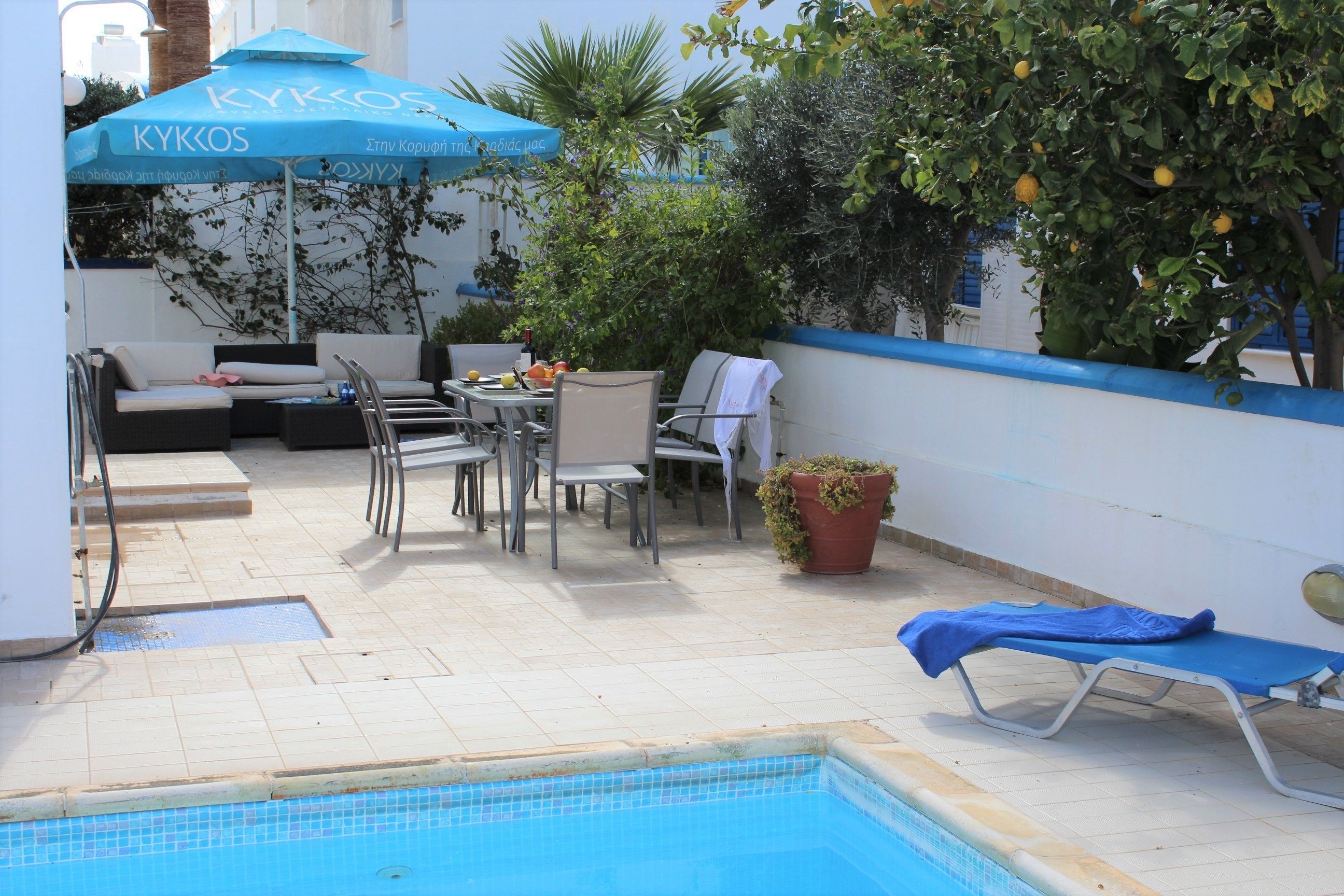 Área da piscina com guarda-sol azul, espreguiçadeiras e uma mesa com frutas.