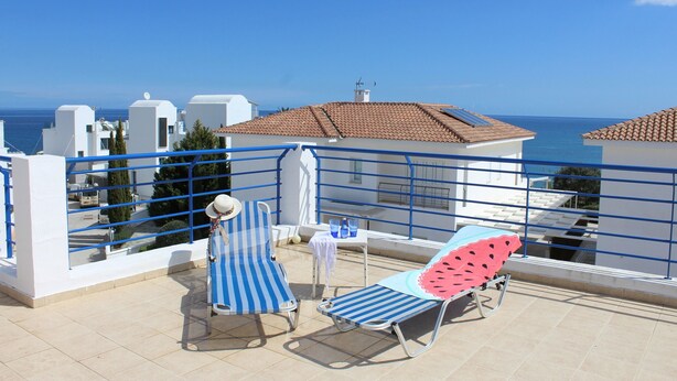 A balcony with two striped lounge chairs and a table, overlooking a coastal area with buildings and the sea. - Sirina Villa STN 29 (Paralimni)