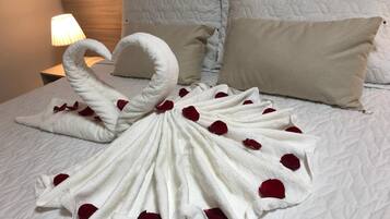 Uma cama bem arrumada com uma toalha disposta em formato de cisne, decorada com pétalas de rosa, e uma luminária na mesa de cabeceira.