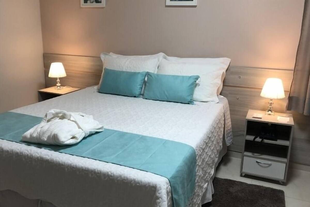 Una cama cuidadosamente hecha con colcha blanca y azul turquesa, dos almohadas a juego y una toalla blanca sobre la cama. Hay dos mesitas de noche con lámparas, una a cada lado de la cama.