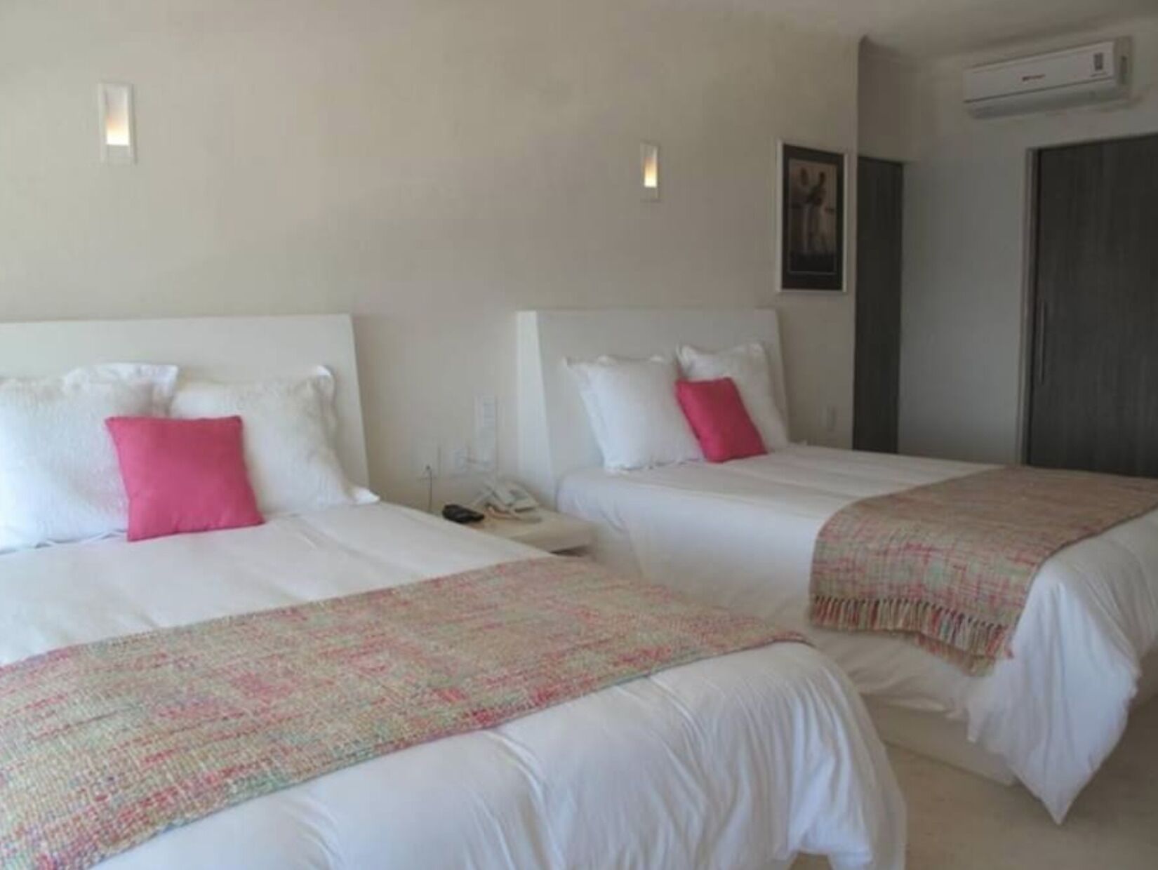 A hotel room with two beds, each with white bedding and a pink pillow. There is a small bedside table with a phone and a lamp. The room has a neutral color scheme with beige walls and a gray door.