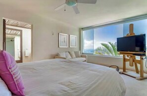 Ein Schlafzimmer mit einem großen Bett, einem Fernseher auf einem Holzständer und einem Fenster mit Blick auf Palmen.