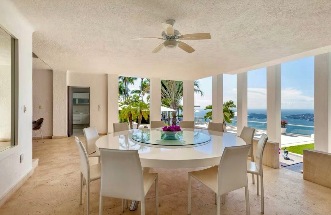A modern dining area with a glass table and white chairs, overlooking a scenic ocean view.