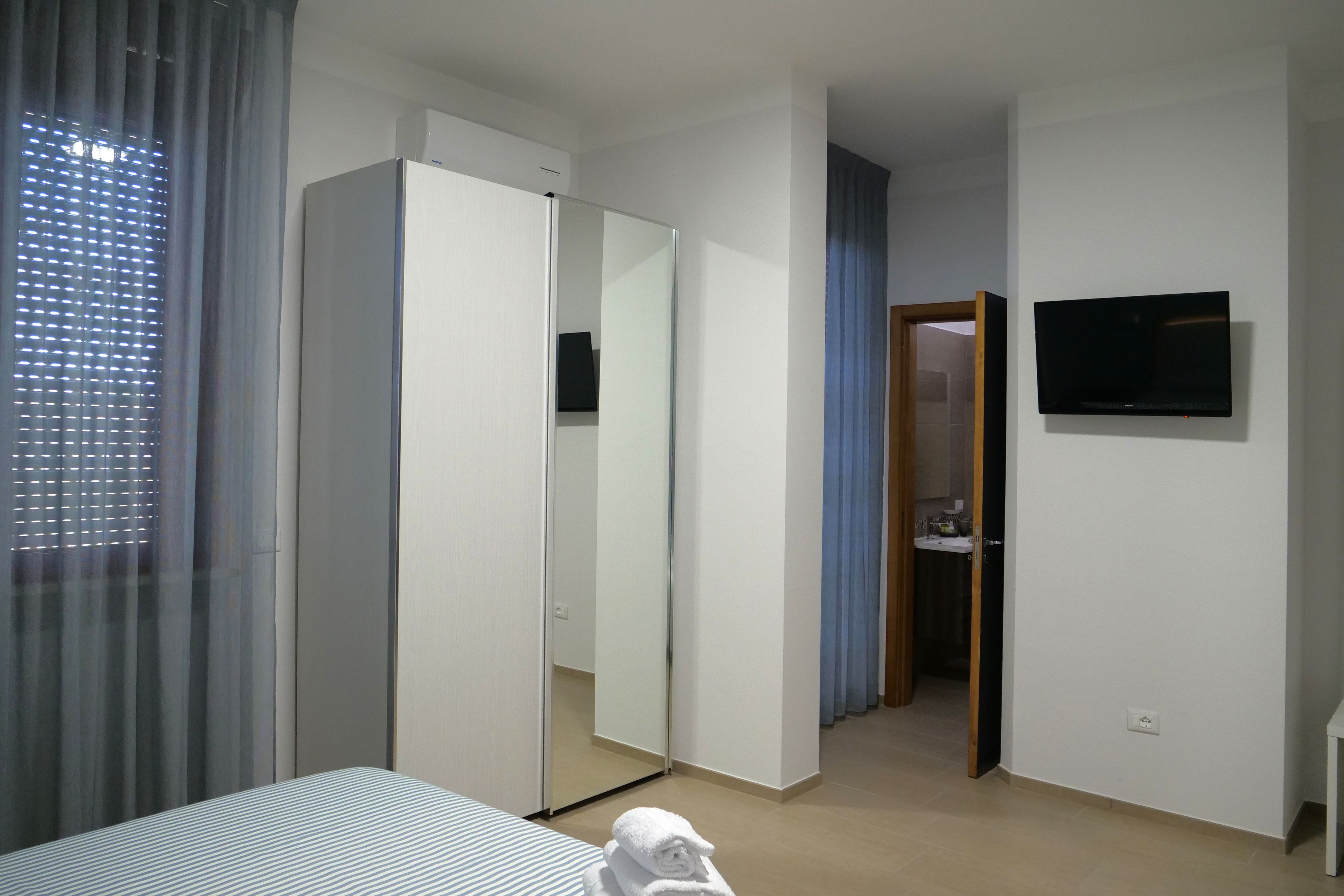 Egy szállodai szoba, amelyben található egy ágy, egy szekrény, egy tükör, egy televízió, és egy fürdőszoba, mely utóbbi egy nyitott ajtón keresztül látható.